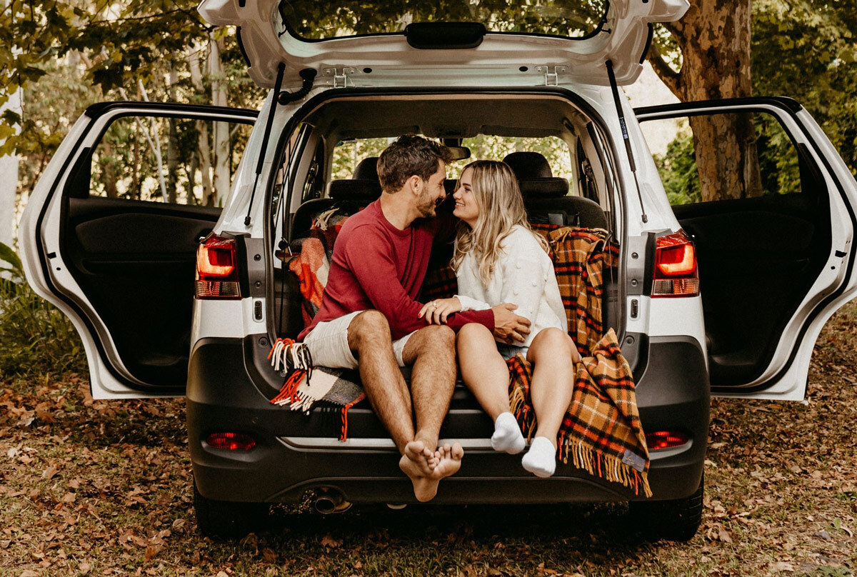 A man and woman sitting in the open trunk of their used vehicle. 